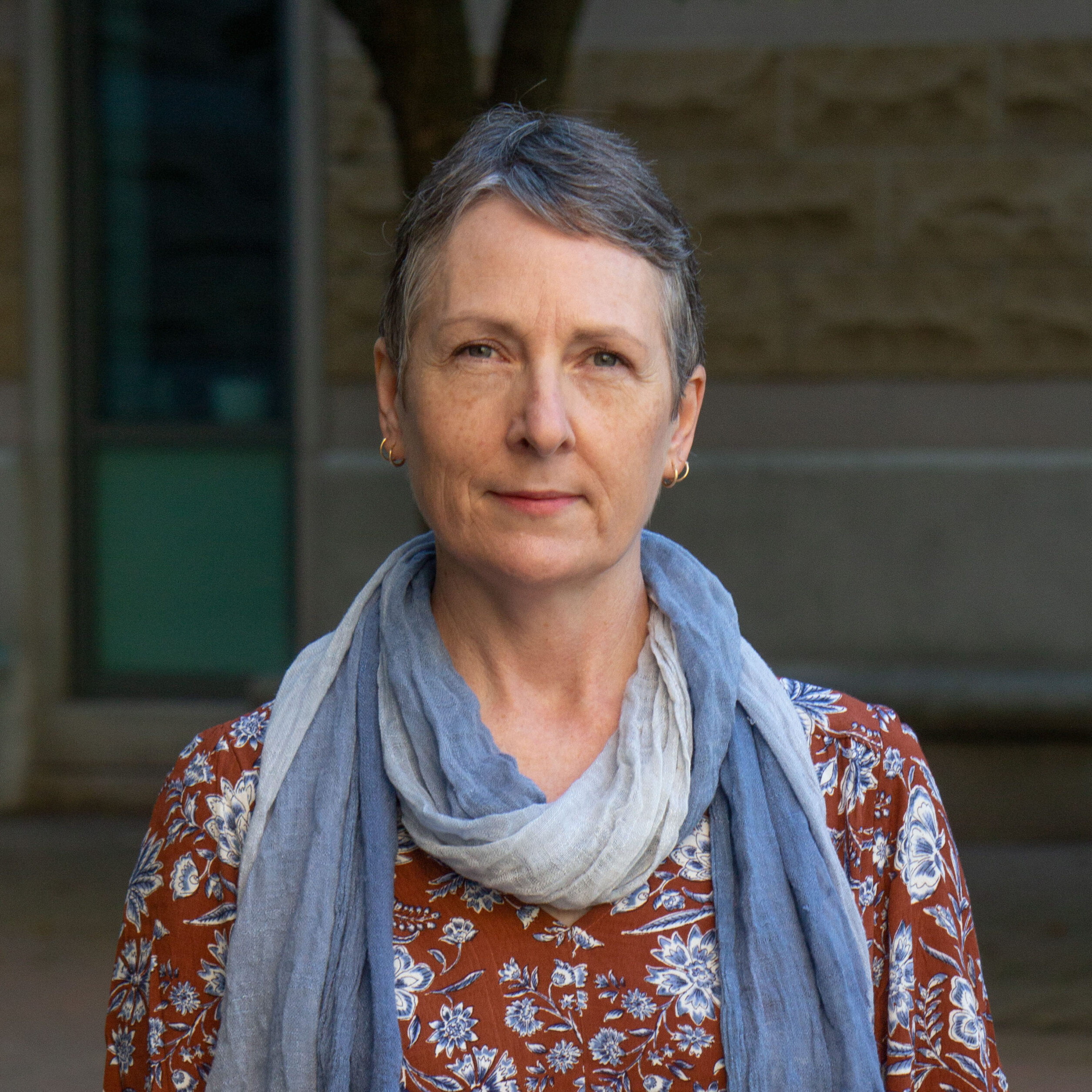 Photo d'une personne aux cheveux courts, portant un foulard bleu et une chemise rouge et bleue, debout devant un fond sombre et délavé.