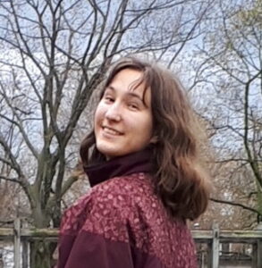 Photo d'une personne aux cheveux bruns vêtue d'une veste rouge, debout devant des arbres.