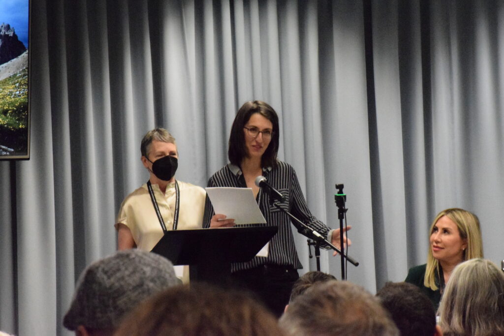 Two white, femme-presenging individuals speaking to an audience at the front of room with a black podium/stand in front of them.