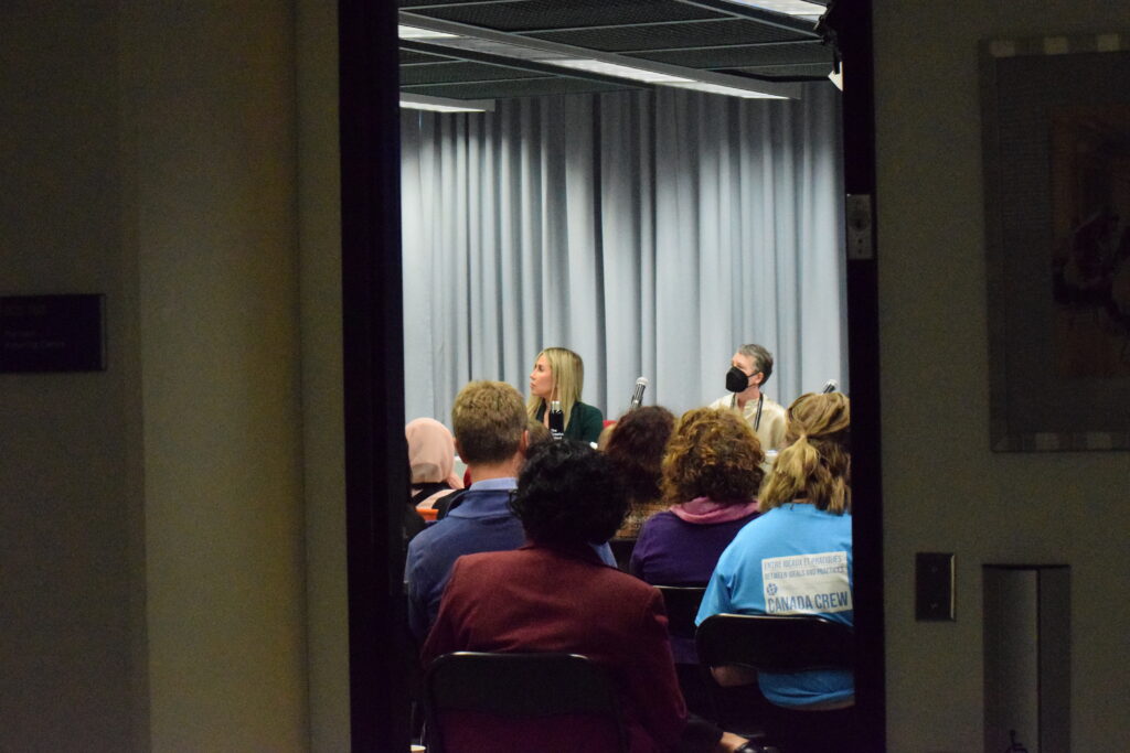 A photograph peaking through the door into a group of people, seated, listening to a presentation