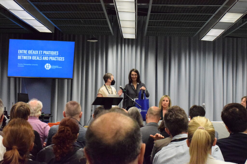 Two people at the front of a room presenting in front of group of individuals. To their left is a blue screen that reads: "Entre Ideaux et Pratiques" "Between Ideals and Practices"
