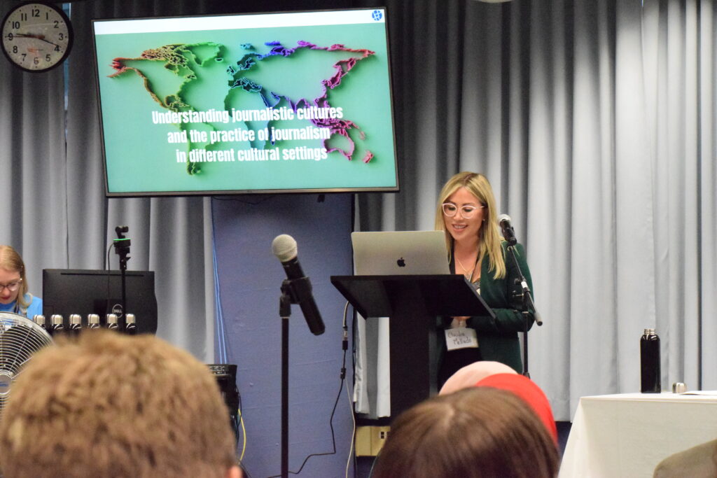 A femme presenting individual with blonde hair smiling, standing in front of a podium with a laptop screen. Behind them is a powerpoint presentation with a title slide that reads: "Understanding Cultures and the Practice of Journalism in different cultural settings