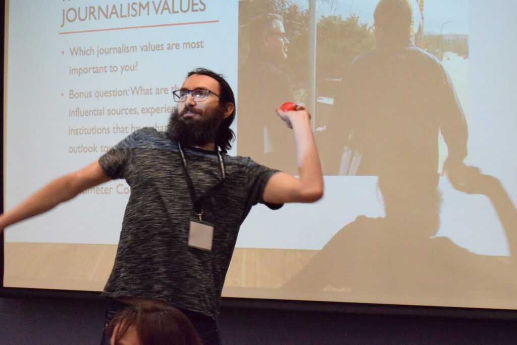 A photograph a masc-presenting individual with long tied-up hair and a long beard. He is holding a red ball and is in the middle of throwing it. He is standing in front of a screen projecting a powerpoint presentation with a slide title that reads: "Journalism Values."