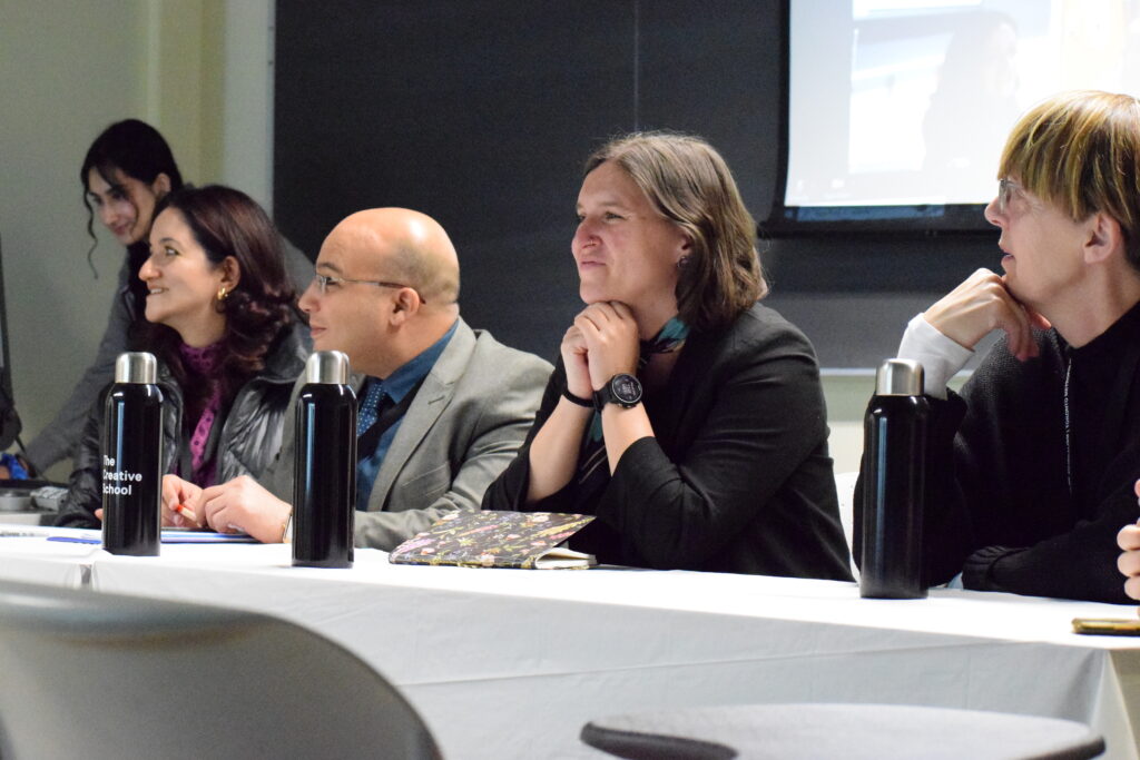 Four individuals sit smiling on a panel. In front of each of them, on the table, is a black stainless steal waterbottle with the words The Creative School.