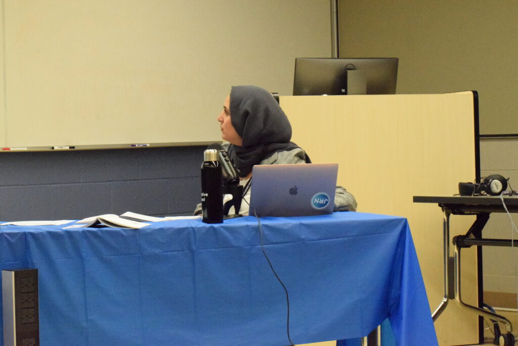 A hijabi femme presenting individual is seated in on a table with a blue table cloth and looks to a presentation in front of them