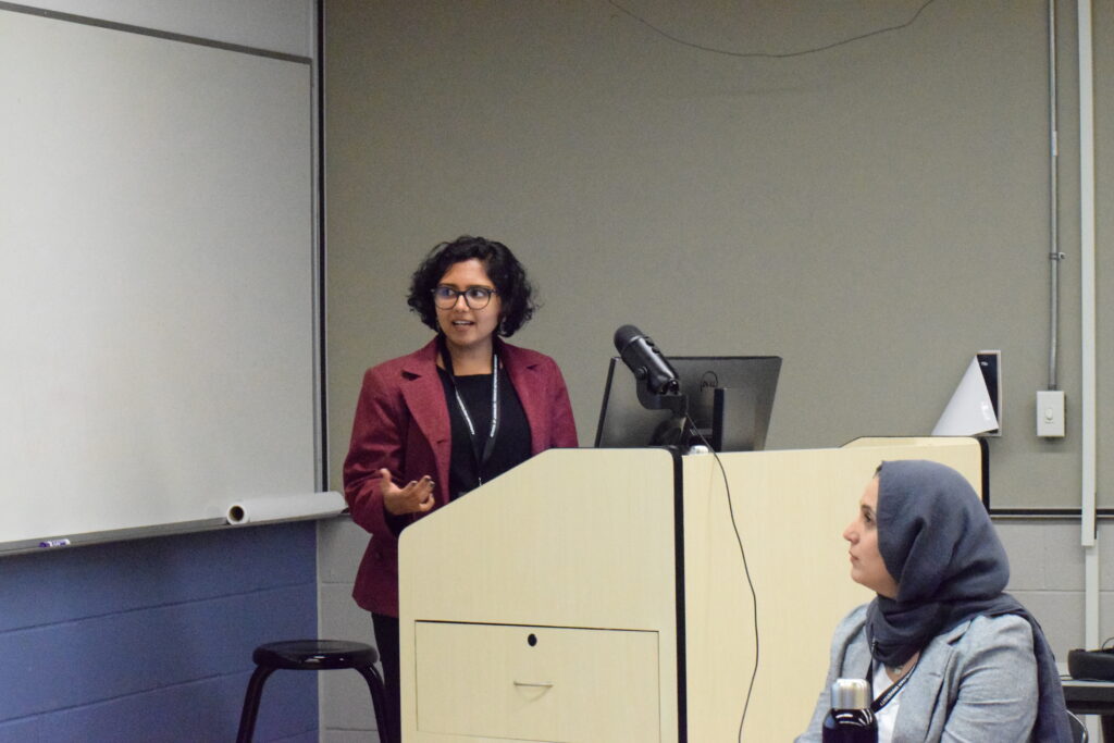 A femme-presenting individual with brown skin and short black hair stands in front of podium. They are looking to their presentation. In the foreground if a hijabi femme presenting individual looking to the presentation.