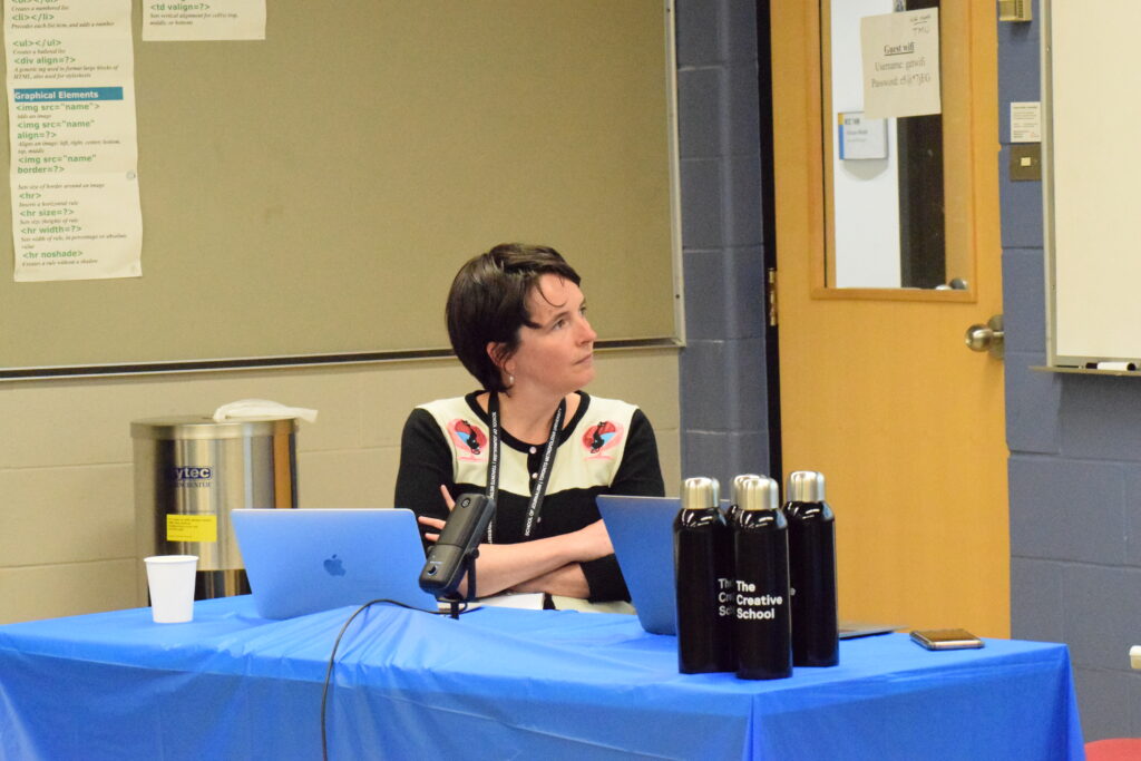 A photograph of a white femme-presenting person with a pixie cut sitting with their arms crossed staring at a screen. The table in front of them has a blue tablecloth on it and four black stainless steal water bottles.