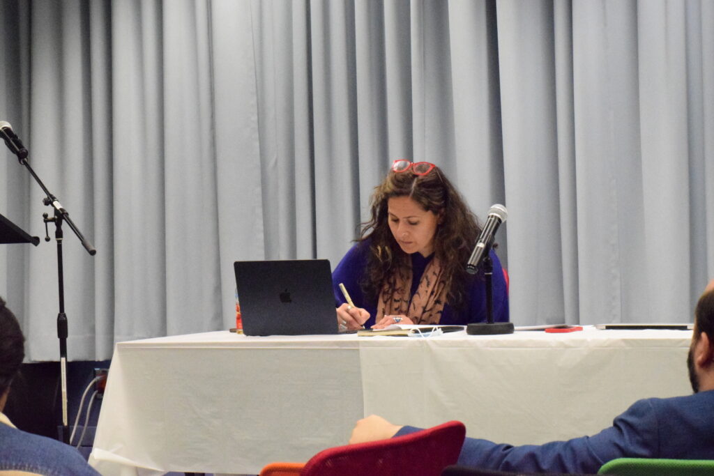 A photograph of a femme presenting individual sitting at a panel table with a microphone and a laptop in front of them.