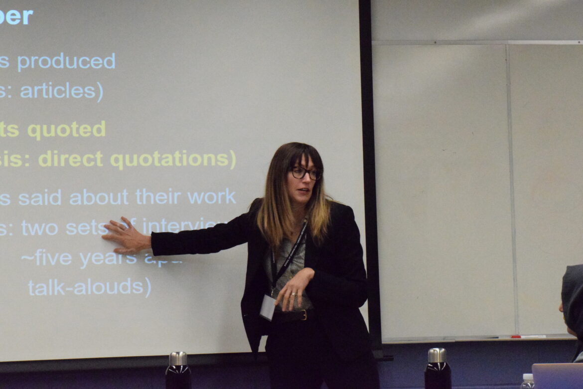 DSC_1576 A white femme presenting individual with long hair and bangs, standing and touching a projection of a presentation on screen.