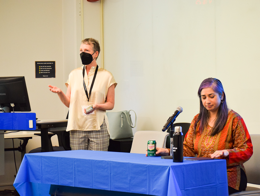 A white femme-presenting individual wearing a black KN95 mask speaks to an audience while standing at the front of a room. Next to them is an individual with long purple hair looking down on a table with a blue table cloth.