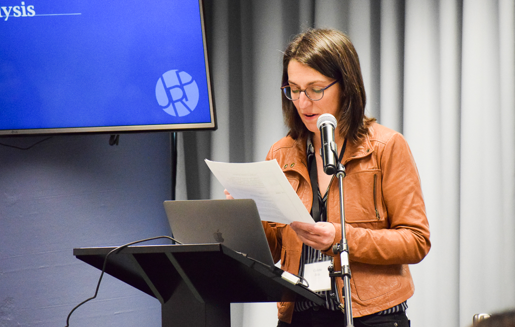 A white femme-presenting person with brown hair, wearing a brown leather jacket, speaks into a microphone on a stand in front of a podium while reading off a script.