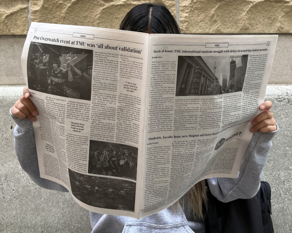 Person with black hair, holding open newspaper that covers face, seated in front of brick wall
