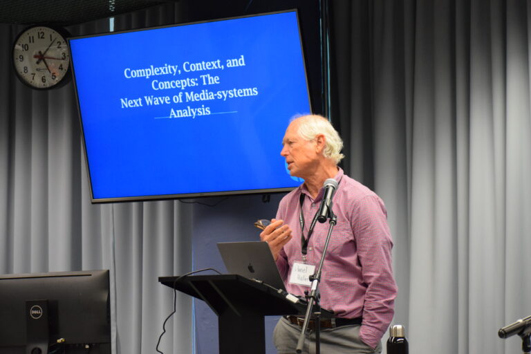 Daniel Hallin stands behind a small, black podium with a large. blue screen behind him that reads "Complexity, context, and concepts: The next wave of media-systems analysis." He has grey hair and is wearing a checkered red long-sleeved shirt and grey pants, and is gesturing with his right arm.