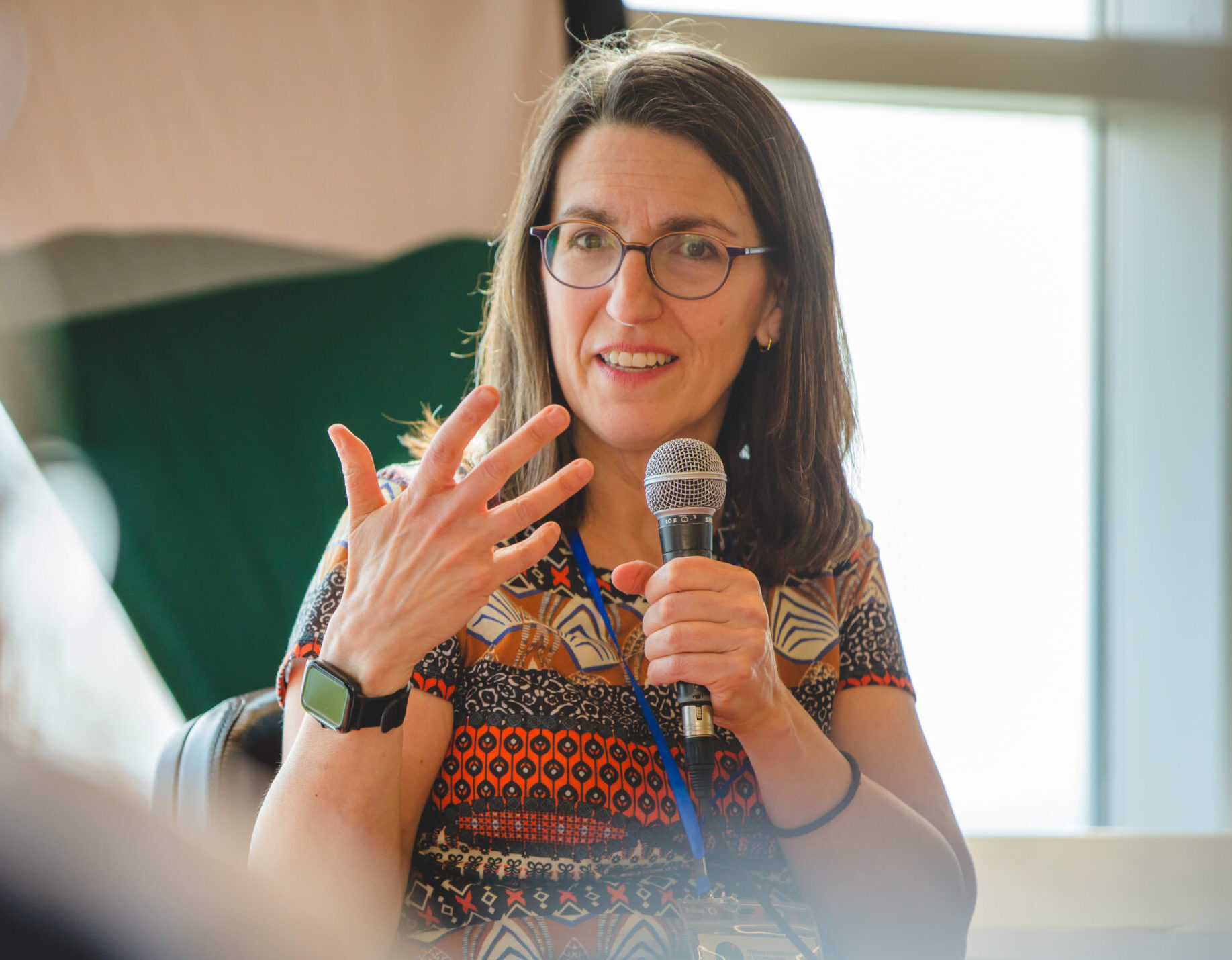 A photo of a person with short brown hair wearing eyeglasses and a colourful shirt holding a microphone.