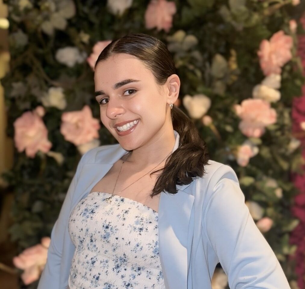 A photo of a person with brown hair in a ponytail wearing a baby blue blazer and a floral top standing in front of blurred background with pink florals.