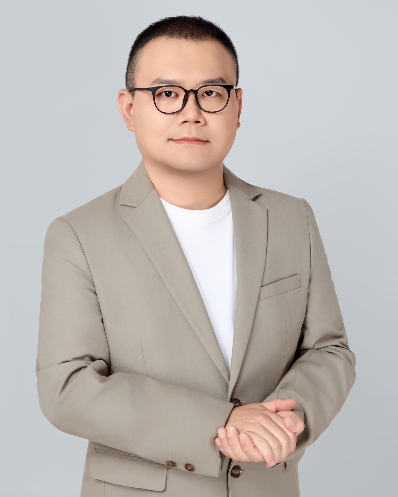 A photo of a person in a tan suit wearing black glasses in front of a light grey background.