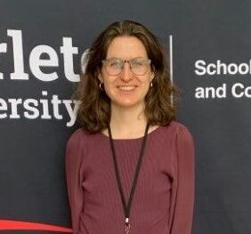 A photo of a woman with brown hair wearing a burgundy shirt and grey pants standing in front of a black and red backgound.