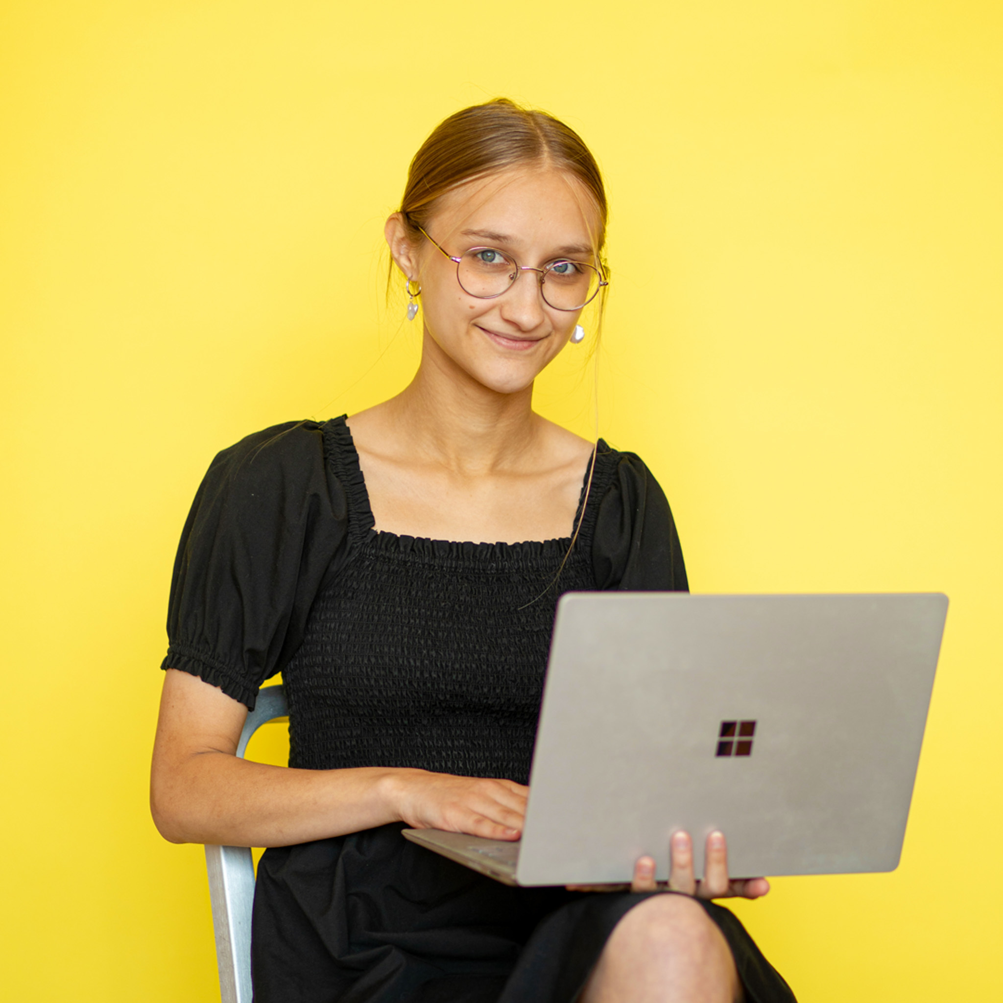 A photo of a woman with dark blonde hair wearing glasses and a black dress holding a computer while sitting in front of a yellow background.