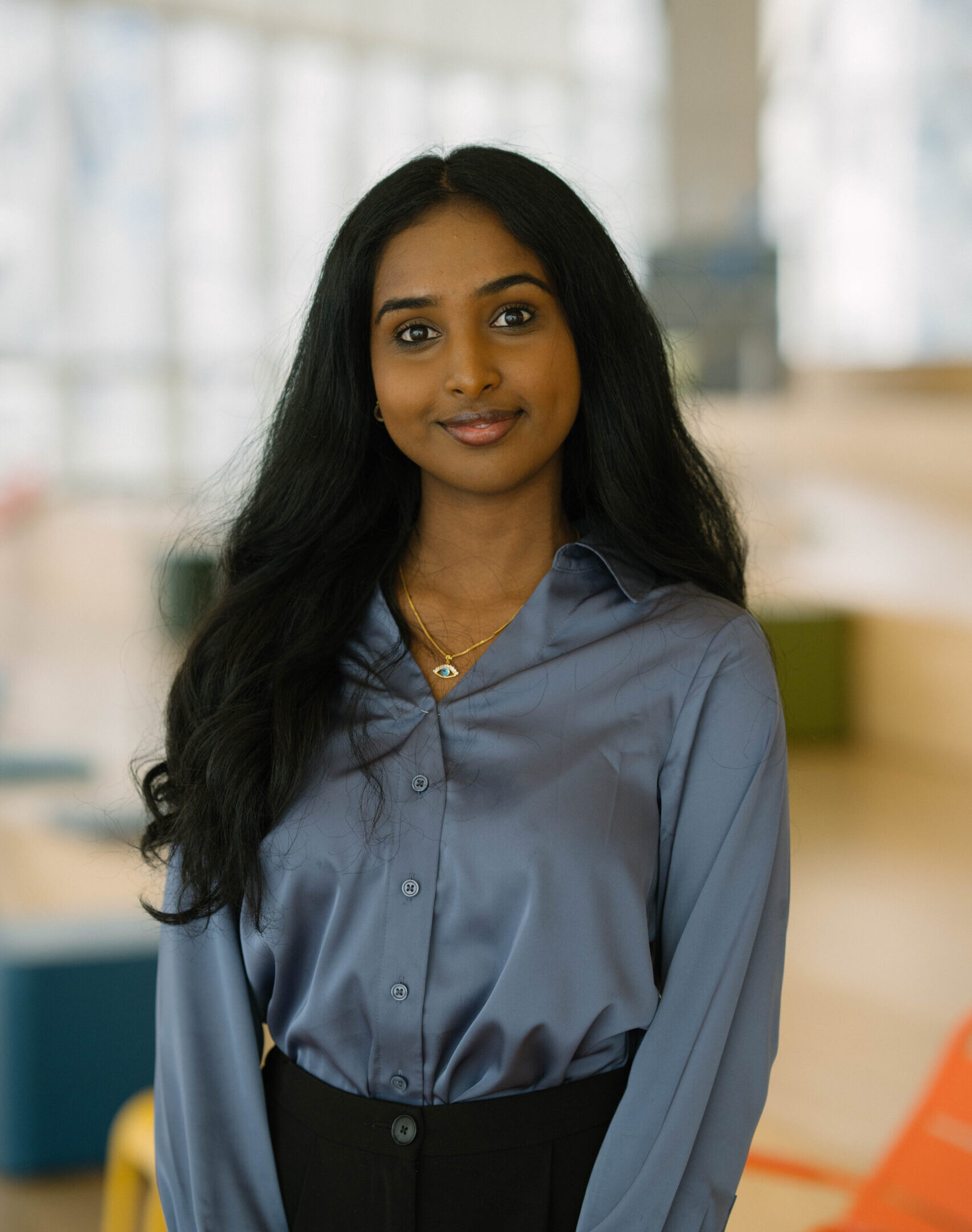 A woman with long black hair wearing a light blue blouse and black pants standing in front of a blurred background